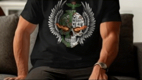 A person wearing a black t-shirt with a skull design and a winged emblem is sitting on a couch.