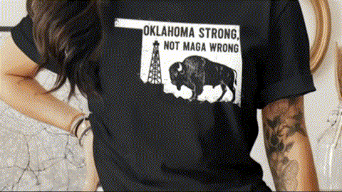 A person wearing a black t-shirt with a graphic design featuring a bison and a tower, along with the text "OKLAHOMA STRONG, NOT MAGA WRONG".