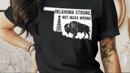 A person wearing a black t-shirt with a graphic design featuring a bison and a tower, along with the text "OKLAHOMA STRONG, NOT MAGA WRONG".