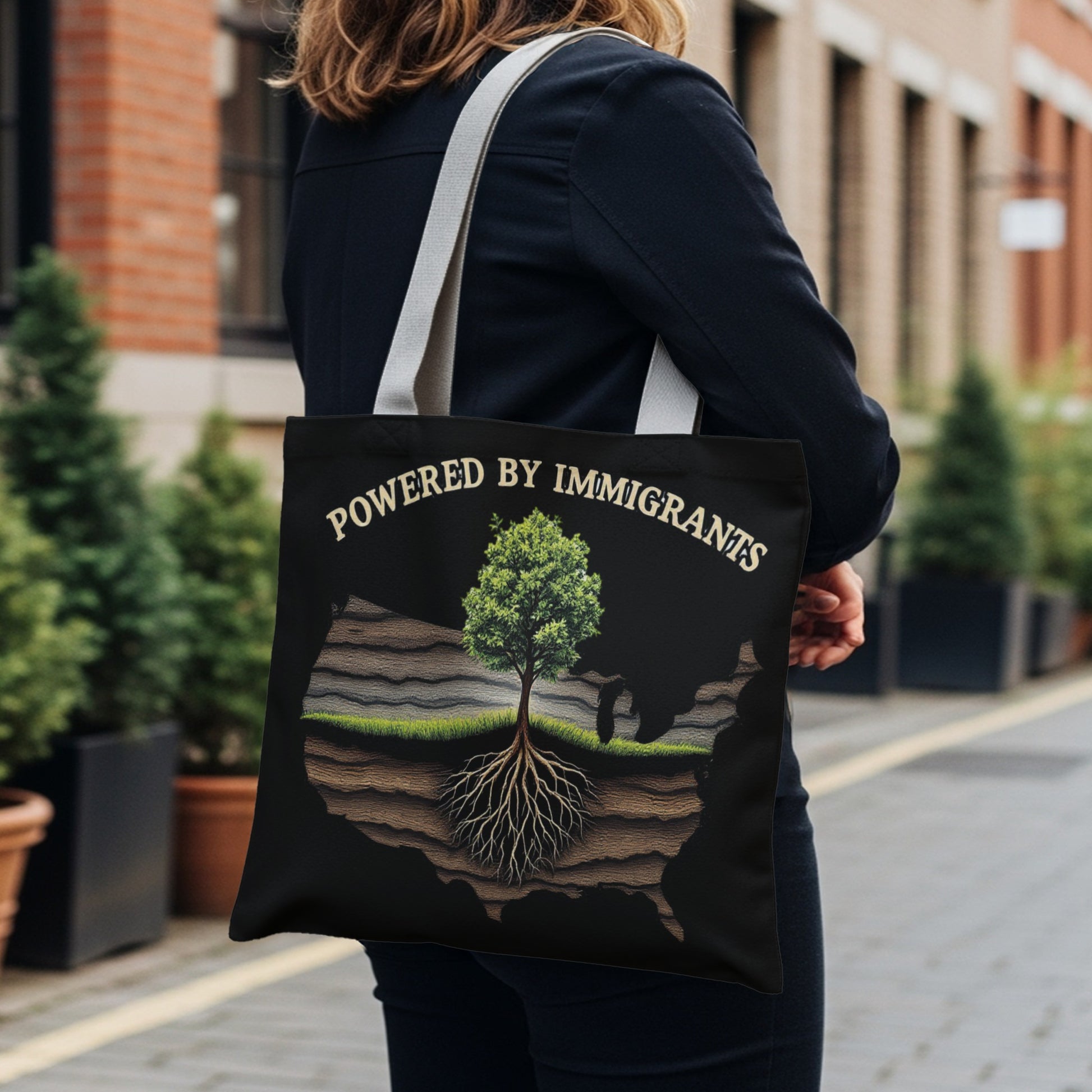 Black tote bag with "Powered by Immigrants" text and a tree growing from a cross-section of land within a US map outline.