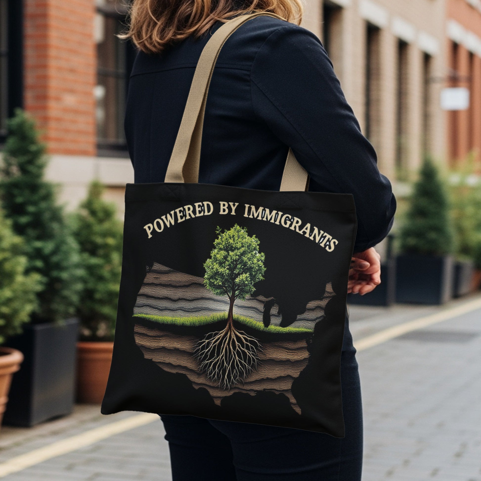 Black tote bag with "POWERED BY IMMIGRANTS" and tree with roots over US map.