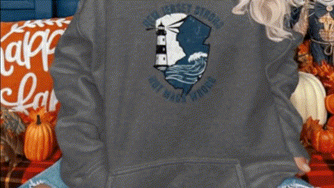 A person wearing a gray sweatshirt with a lighthouse design on it, sitting on a couch surrounded by fall decorations.