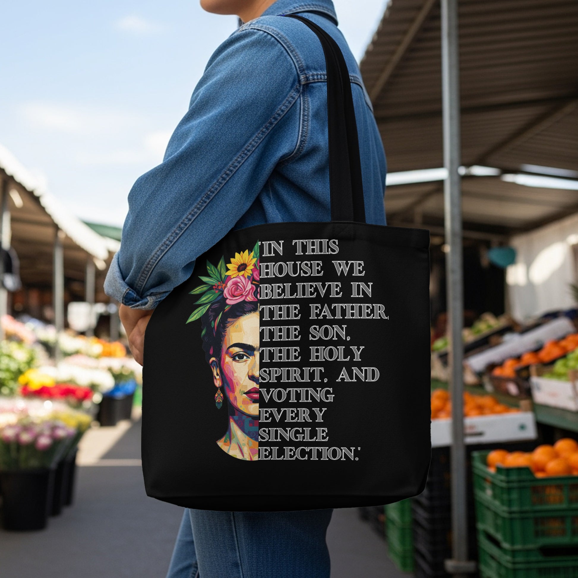 Black tote bag with text "In this house we believe in the Father, the Son, the Holy Spirit, and voting every single election" and a portrait of Frida Kahlo.