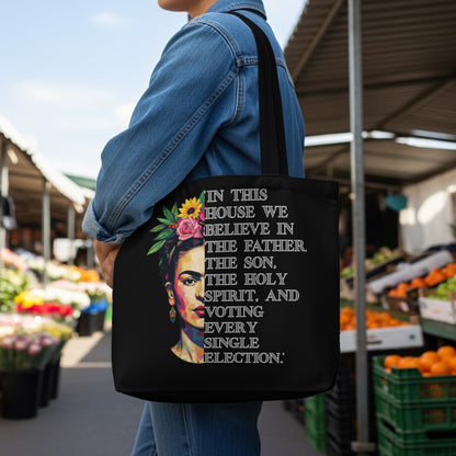 Black tote bag with text "In this house we believe in the Father, the Son, the Holy Spirit, and voting every single election" and a portrait of Frida Kahlo.